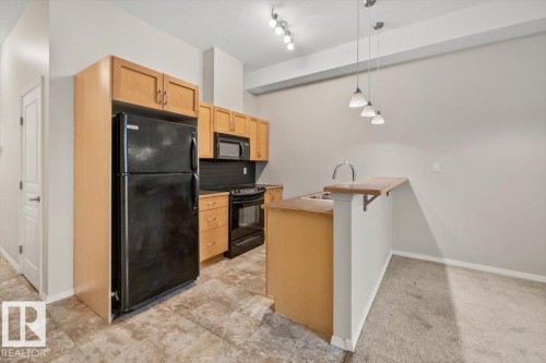 Kitchen featuring wood-finish cabinetry, black appliances, track lighting, and a peninsula with pendant lighting and bar seating - 228 10531 117 Street, Edmonton, AB - Indoor Photo Showing Kitchen