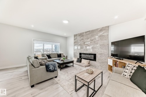 Living room featuring light-toned flooring, a light-colored area rug, a fireplace with a stone surround, and large windows providing views of the outdoors - 4509 170A Avenue, Edmonton, AB - Indoor Photo Showing Living Room With Fireplace