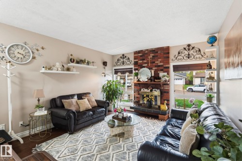 Living area featuring a brick fireplace with a brass insert, large front-facing windows, built-in shelving, hardwood flooring, and a textured ceiling - 15232 120 Nw, Edmonton, AB 
