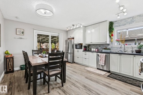 Modern kitchen and dining area featuring light wood-finish flooring, white cabinetry, stainless steel appliances, and dark countertops - 15232 120 Nw, Edmonton, AB 