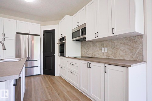 Contemporary kitchen featuring white shaker cabinetry, stainless steel appliances, a light-toned backsplash, light wood-finish flooring, and dark countertops - 82 Meadowview Point(E), Sherwood Park, AB - Indoor Photo Showing Kitchen