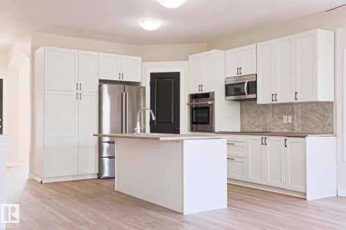 Modern kitchen featuring a central island with an integrated sink, stainless steel appliances, white shaker-style cabinetry, light wood-finish flooring, and a stone backsplash - 82 Meadowview Point(E), Sherwood Park, AB - Indoor Photo Showing Kitchen With Upgraded Kitchen