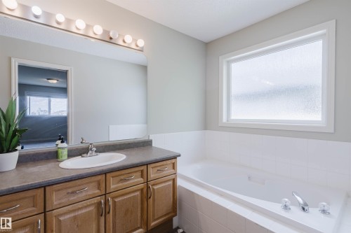 Bathroom featuring a built-in tub with tile surround, a large vanity mirror with strip lighting, a single vanity with storage cabinetry, and a frosted glass window - 3534 Mclean Crescent, Edmonton, AB - Indoor Photo Showing Bathroom