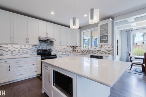 Kitchen featuring a large central island with a built-in microwave, white shaker-style cabinetry, mosaic tile backsplash, and wood-finish flooring - 3571 Keswick Boulevard, Edmonton, AB - Indoor Photo Showing Kitchen With Upgraded Kitchen