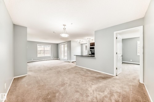 The open living area features neutral-toned carpeting, light grey walls, and a large window with white framing - 101 530 Watt Boulevard, Edmonton, AB - Indoor Photo Showing Other Room