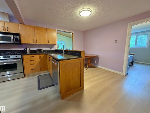 Kitchen featuring light wood-finish flooring, wood cabinetry, stainless steel appliances, black countertops, and a tile backsplash - 60016 Rr 41, Rural Barrhead County, AB - Indoor