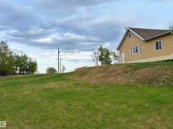 Expansive grassy terrain with a gentle slope leading to a residence featuring a light yellow siding exterior, white trim, and a brown shingle roof - 