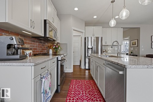 Modern kitchen featuring white shaker cabinetry, stainless steel appliances, a center island with sink, exposed brick backsplash, and wood-finish flooring - 1359 Westerra Drive, Stony Plain, AB - Indoor Photo Showing Kitchen With Upgraded Kitchen