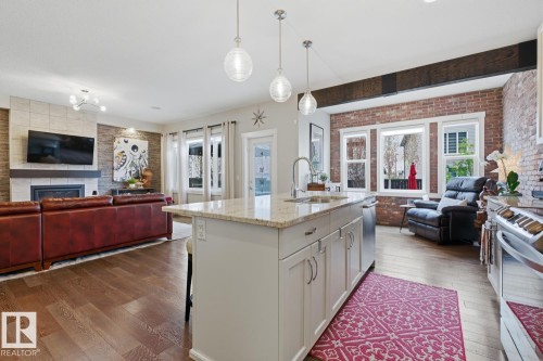 Open-concept living space featuring a kitchen island with stone countertop, wood-finish flooring, a contemporary fireplace with a tiled surround, exposed brick accent wall, and recessed and pendant lighting - 1359 Westerra Drive, Stony Plain, AB - Indoor With Fireplace