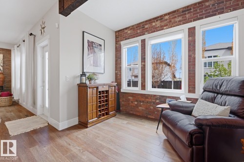 Living area featuring an exposed brick accent wall, white trim windows, wood-finish flooring, a white paneled door, and a prominent exposed ceiling beam - 1359 Westerra Drive, Stony Plain, AB - Indoor