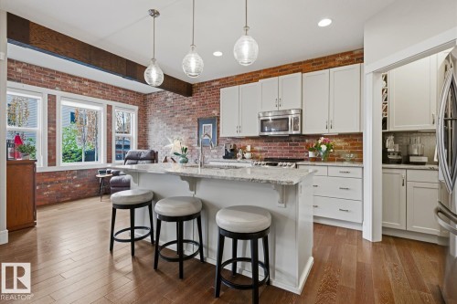 Open-concept kitchen featuring an oversized island with countertop seating, wood-finish flooring, exposed brick accents, white cabinetry, and stainless steel appliances - 1359 Westerra Drive, Stony Plain, AB - Indoor Photo Showing Kitchen With Upgraded Kitchen