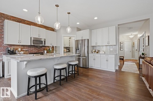 Open-concept kitchen featuring wood-finish flooring, white cabinetry, stainless steel appliances, a breakfast bar with pendant lighting, and a brick accent wall - 1359 Westerra Drive, Stony Plain, AB - Indoor Photo Showing Kitchen With Upgraded Kitchen