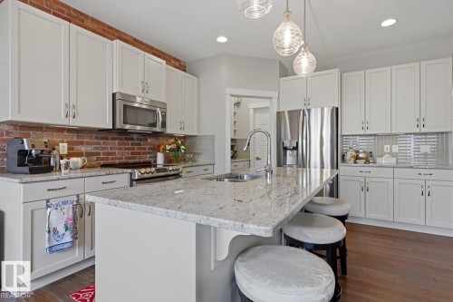 Spacious kitchen featuring an expansive central island with a sink, stainless steel appliances, white shaker cabinetry, a brick backsplash, and wood-finish flooring - 1359 Westerra Drive, Stony Plain, AB - Indoor Photo Showing Kitchen With Double Sink With Upgraded Kitchen