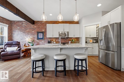 Open-concept kitchen featuring an eat-at island with pendant lighting, stainless steel appliances, brick accent wall, wood-finish flooring, and white cabinetry - 1359 Westerra Drive, Stony Plain, AB - Indoor Photo Showing Kitchen With Upgraded Kitchen