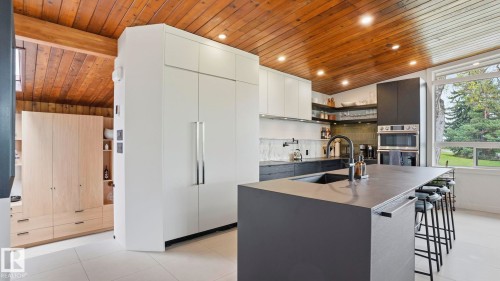 Modern kitchen featuring a large central island with integrated sink, sleek white and dark cabinetry, exposed wood-paneled vaulted ceiling, built-in double ovens, and a large window providing ample natural light - 6412 Ada Boulevard, Edmonton, AB - Indoor Photo Showing Kitchen