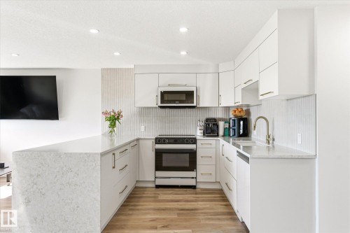 Modern kitchen featuring white cabinetry with gold-tone hardware, a U-shaped countertop with a waterfall edge, stainless steel appliances, a white vertical tile backsplash, and wood-finish flooring - 108 10105 95 Street, Edmonton, AB - Indoor Photo Showing Kitchen With Upgraded Kitchen