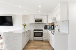 Modern kitchen featuring white cabinetry with gold-tone hardware, a U-shaped countertop with a waterfall edge, stainless steel appliances, a white vertical tile backsplash, and wood-finish flooring - 