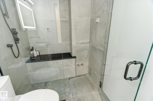 Contemporary shower enclosure featuring full-height glass panels, matte black shower fixtures, a built-in bench with a dark countertop, and large format gray tile surrounds - 108 10105 95 Street, Edmonton, AB - Indoor Photo Showing Bathroom