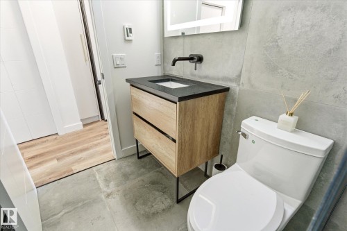 Modern bathroom featuring a wood-finish vanity with a black countertop, a wall-mounted matte black faucet, and a large illuminated mirror - 108 10105 95 Street, Edmonton, AB - Indoor Photo Showing Bathroom