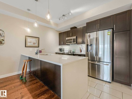 Contemporary kitchen featuring stainless steel appliances, dark wood-finish cabinetry, a white countertop island, and pendant lighting - 213 625 Leger Way, Edmonton, AB - Indoor Photo Showing Kitchen With Stainless Steel Kitchen