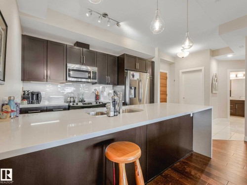Modern kitchen with a large central island, white countertop, dark wood-finish cabinetry, stainless steel appliances, and wood-finish flooring - 213 625 Leger Way, Edmonton, AB - Indoor Photo Showing Kitchen With Stainless Steel Kitchen With Double Sink With Upgraded Kitchen