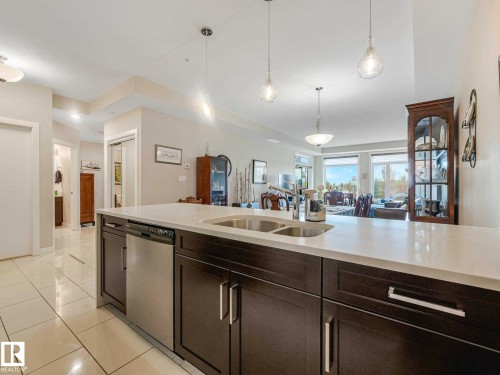 Kitchen island featuring a dual-basin stainless steel sink, integrated dishwasher, and dark wood-finish cabinetry with silver-tone hardware - 213 625 Leger Way, Edmonton, AB - Indoor Photo Showing Kitchen With Double Sink