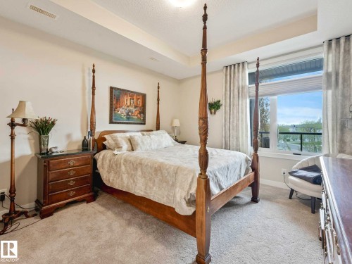 Bedroom featuring a large window, light-colored carpeting, a tray ceiling with recessed lighting, and neutral wall tones - 213 625 Leger Way, Edmonton, AB - Indoor Photo Showing Bedroom