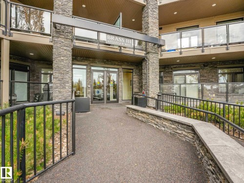Stone facade entry featuring a covered entrance, glass doors, black railings, and a ramp for accessibility - 213 625 Leger Way, Edmonton, AB - Outdoor With Balcony With Exterior