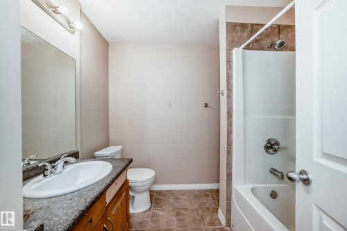 Bathroom featuring a white bathtub and shower combination with brown tile surround, a built-in wood-finish vanity with a dark countertop and undermount sink, and tiled flooring - 9825 156 Street, Edmonton, AB - Indoor Photo Showing Bathroom