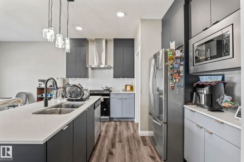 Modern kitchen featuring a spacious island with an undermount sink, pendant lighting, stainless steel appliances, dark cabinetry, and wood-finish flooring - 5142 23A Avenue, Edmonton, AB - Indoor Photo Showing Kitchen With Double Sink With Upgraded Kitchen