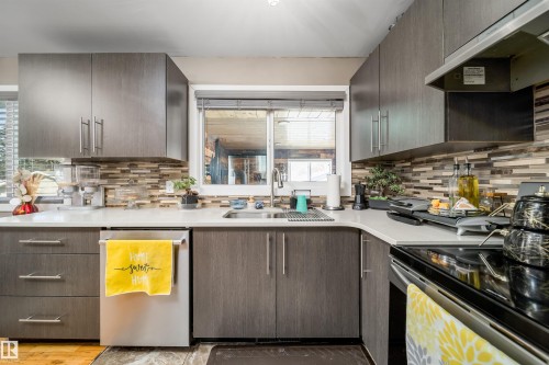 Contemporary kitchen featuring wood-grain cabinetry with brushed nickel hardware, light-toned countertops, a glass tile backsplash, and stainless steel appliances including a dishwasher and range - 14808 96 Street, Edmonton, AB - Indoor Photo Showing Kitchen With Upgraded Kitchen