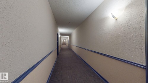 Long interior corridor featuring wall-mounted lighting fixtures, textured walls with a two-tone paint scheme, and a patterned carpet runner - 405 16303 95 Street Nw, Edmonton, AB - Indoor Photo Showing Other Room