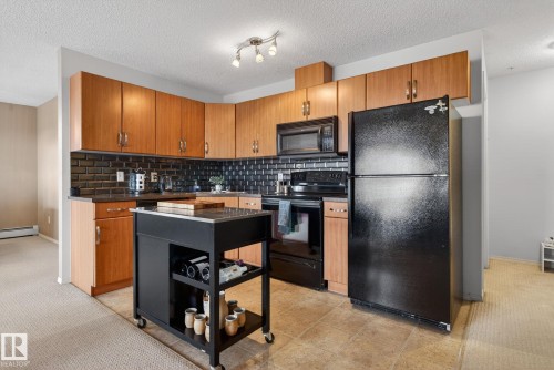 The kitchen features wood cabinetry, dark countertops, a black subway tile backsplash, and a black refrigerator - 423 2430 Guardian Road, Edmonton, AB - Indoor Photo Showing Kitchen