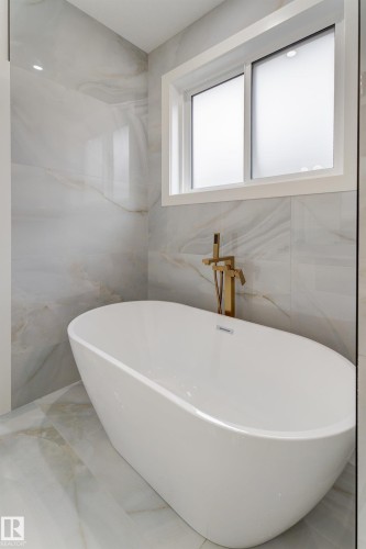 Freestanding soaking tub with a floor-mounted gold-tone faucet, set against large format wall and floor tiles featuring a subtle veined pattern - 4727 17 Avenue, Edmonton, AB 