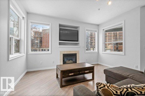 Living area featuring wood-finish flooring, light gray walls, a built-in fireplace with a tiled surround, a recessed entertainment niche, and multiple windows with white trim - 304 10011 110 Street, Edmonton, AB - Indoor Photo Showing Living Room With Fireplace