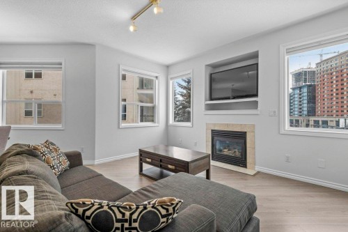 Living area featuring light wood-finish flooring, multiple windows, a built-in media niche, and a fireplace with a stone surround and hearth - 304 10011 110 Street, Edmonton, AB - Indoor Photo Showing Living Room With Fireplace