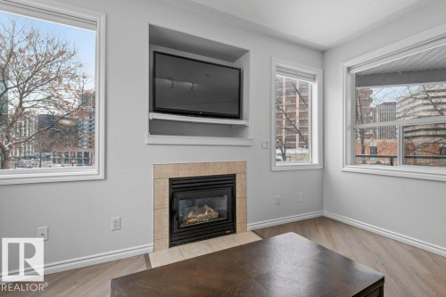 Living area featuring a tiled fireplace with an integrated media niche, wood-finish flooring, and multiple windows - 304 10011 110 Street, Edmonton, AB - Indoor Photo Showing Living Room With Fireplace