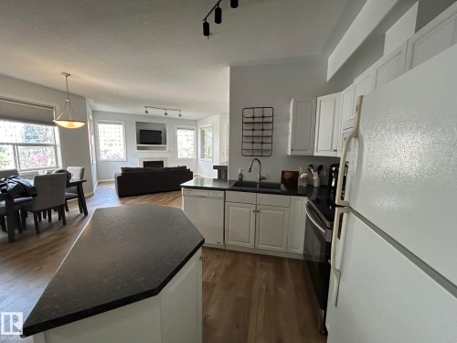 Open-concept kitchen featuring white cabinetry, dark countertops, built-in dishwasher, and wood-finish flooring extending into living and dining areas - 304 10011 110 Street, Edmonton, AB - Indoor Photo Showing Kitchen