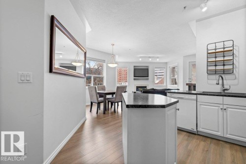 Open-concept living space featuring a kitchen island with dark countertops, white cabinetry, wood-finish flooring, a dining area with pendant lighting, and a living area with track lighting - 304 10011 110 Street, Edmonton, AB - Indoor Photo Showing Kitchen