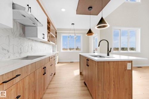 Spacious kitchen featuring a central island with an undermount sink and matte black faucet, wood-finish cabinetry, white upper cabinets, hexagonal tile backsplash, and light wood-finish flooring - 4915 17 Avenue, Edmonton, AB - Indoor Photo Showing Kitchen With Double Sink With Upgraded Kitchen