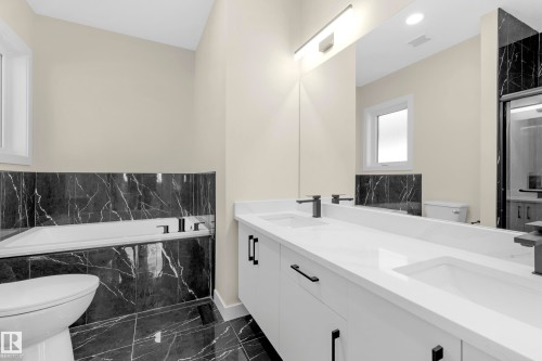 Bathroom featuring a dual vanity with white countertops and integrated sinks, matte black hardware, a built-in tub with black marble-effect tile surround, and coordinating black marble-effect floor tiling - 4915 17 Avenue, Edmonton, AB - Indoor Photo Showing Bathroom