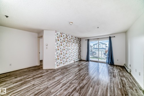Spacious room featuring wood-finish flooring, a patterned accent wall, and a sliding glass door with dark blue drapes - 210 2305 35A Avenue, Edmonton, AB - Indoor Photo Showing Other Room