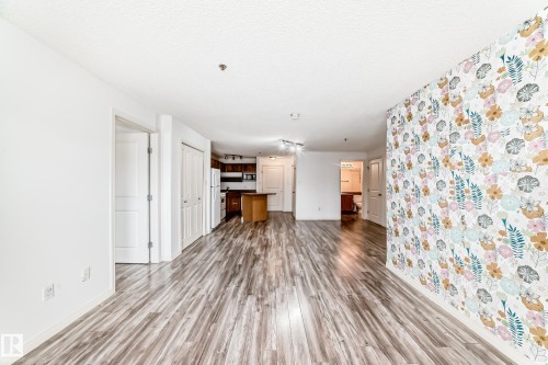 Open-concept living area featuring wood-finish flooring, a decorative accent wall, a track lighting fixture, and a partial view of the kitchen with dark cabinetry and a central island - 210 2305 35A Avenue, Edmonton, AB - Indoor Photo Showing Other Room
