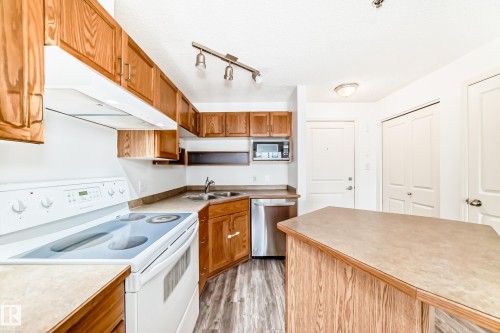 Kitchen featuring wood cabinetry, white appliances, a stainless steel dishwasher, and wood-finish flooring - 210 2305 35A Avenue, Edmonton, AB - Indoor Photo Showing Kitchen With Double Sink