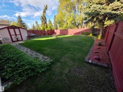 Expansive backyard featuring a manicured lawn, red privacy fencing, a storage shed, a stone paver pathway, and mature trees - 