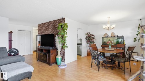 Open-concept living space featuring wood-finish flooring, a dining area with a chandelier, and a partial brick-accent wall - 105 5125 Riverbend Road, Edmonton, AB - Indoor
