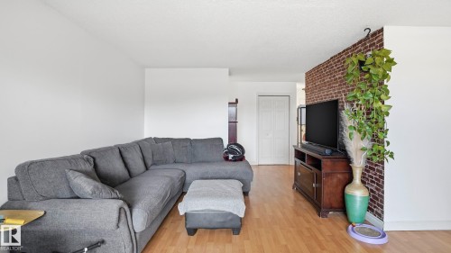Bright living space featuring wood-finish flooring, a white textured ceiling, and a prominent exposed brick accent wall - 105 5125 Riverbend Road, Edmonton, AB - Indoor Photo Showing Living Room