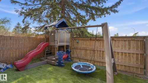 Fenced backyard featuring artificial turf, a children's play structure with a slide and swing set, and a mature evergreen tree providing shade - 105 5125 Riverbend Road, Edmonton, AB - Outdoor