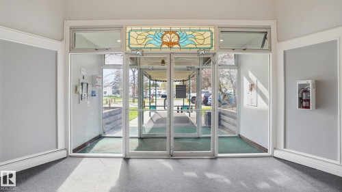 Entrance vestibule featuring a stained glass transom, full-lite glass double doors, a glass sidelight, and a gray carpeted floor - 105 5125 Riverbend Road, Edmonton, AB - Indoor Photo Showing Other Room