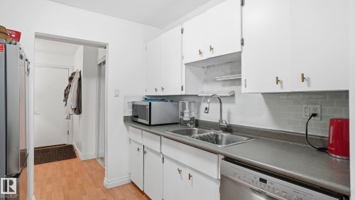 Kitchen featuring white cabinetry with gold-tone hardware, a stainless steel double sink with a gooseneck faucet, gray countertops, and a gray subway tile backsplash - 105 5125 Riverbend Road, Edmonton, AB - Indoor Photo Showing Kitchen With Double Sink